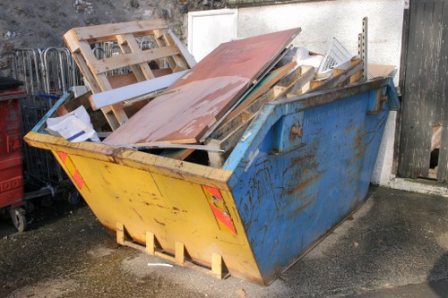 A large metal skip positioned outdoors on a paved surface, painted yellow on the rear and sides with visible rust and dirt, filled with various discarded construction and renovation materials including wooden pallets, MDF boards, metal framing, and cardboard boxes. The skip is situated near a high wall with a weathered wooden fence on the right and a concrete wall behind, indicating a residential or commercial property in the Harrow area. The contents are stacked haphazardly, with some materials leaning against each other and protruding above the skip's top edge. The surrounding environment features dark, damp patches on the ground, consistent with an active waste collection or clearance site. Off to the side, part of another small recycling container or trolley is visible, hinting at a site involved in rubbish removal or clearance services, consistent with the offerings of Office Clearance Harrow.
