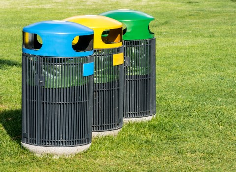 Three cylindrical outdoor rubbish bins are positioned next to each other on a grassy area, with a backdrop of a well-maintained lawn. The bins are made of black plastic with vertical slats, each featuring a colored, rounded top lid: the forefront bin has a blue lid, the middle bin has a yellow lid, and the rear bin has a green lid. Each lid has a smooth, matte finish, with an opening on the side for waste disposal. The outdoor setting appears bright with natural daylight, and there are no other visible objects or structures nearby. These bins are consistent with waste management services such as those offered by Office Clearance Harrow, often used for general rubbish collection and disposal in local areas or commercial premises within Harrow. The placement suggests they are intended for public or employee use on a commercial or communal site, supporting effective rubbish segregation and disposal.