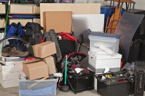 A large collection of various household and office items awaiting disposal, arranged in a cluttered space, possibly a storage area or driveway in Harrow. The foreground features several cardboard boxes, some sealed, along with plastic storage containers, one transparent with neatly folded blue fabric inside. A black toolbox and a set of cleaning tools, including a green mop handle, are visible among the clutter. Leaning against the items are a black backpack and a red-handled tool, with additional objects such as a small stack of papers, a white foam box, and various electronic cables and miscellaneous items scattered throughout. In the background, there are wooden and metal chairs, suggesting a residential or commercial environment undergoing clearance. The scene is well-lit, likely by natural light, emphasizing the mixture of textures from cardboard, plastic, metal, and fabric. This image exemplifies the type of household waste and office items that Office Clearance Harrow would handle as part of their rubbish removal services within the local Harrow area and surrounding postcode district.