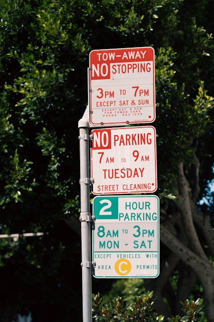 A metal pole standing outdoors against a backdrop of dense green foliage with a clear blue sky visible through the leaves. Attached to the pole are three rectangular traffic and parking restriction si