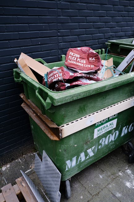 A large green wheeled rubbish skip positioned outdoors on a paved surface next to a dark brick wall. The skip is filled with red and black packaging bags, likely containing construction or garden mate