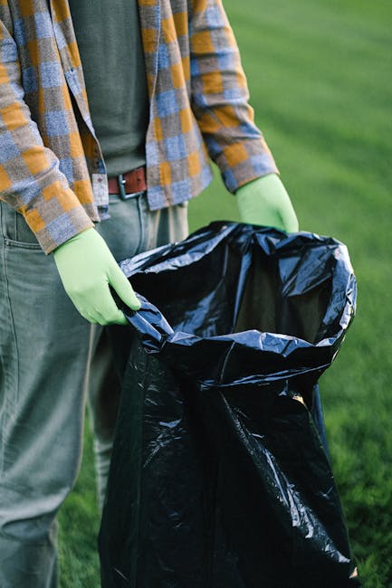A person wearing a multicoloured checkered shirt, grey pants, and bright green gloves is holding open a large black rubbish bag, which appears to be made of durable plastic, in an outdoor setting with
