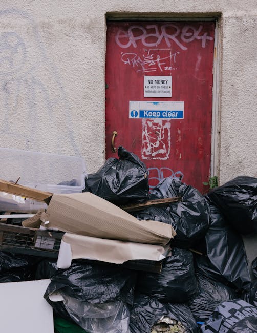 A person wearing a multicoloured checkered shirt, grey pants, and bright green gloves is holding open a large black rubbish bag, which appears to be made of durable plastic, in an outdoor setting with